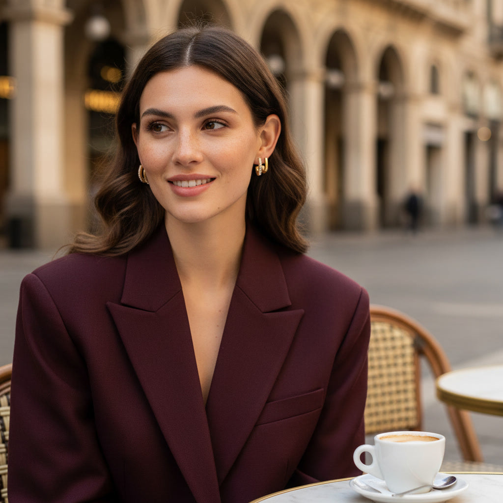Woman in burgundy blazer wearing gold huggie  earrings seated at Milanese outdoor café —  how to style gold earrings by Madam Lust
